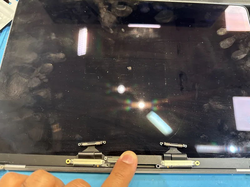 A laptop screen assembly on a blue repair mat, with its hinges and display connector exposed and a finger pointing at the connector