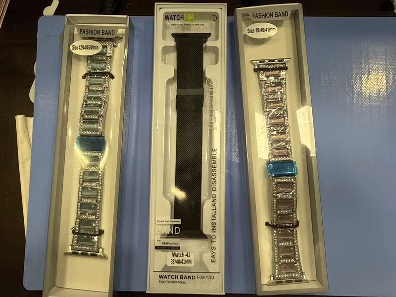 Three boxed smartwatch bands, two jeweled bands and one black mesh band, on a blue mat